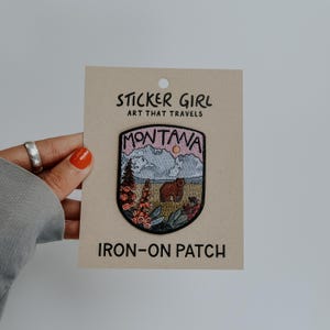 May include: An embroidered iron-on patch on a tan backing card labeled STICKER GIRL ART THAT TRAVELS. The patch features a shield shape with the text MONTANA, a brown bear, a mountain range, a sun, and colorful wildflowers in a landscape.