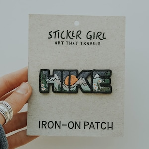May include: Embroidered iron-on patch with the word "HIKE" in a stylized design featuring mountains, a setting sun, and flowers. The patch is on a cardboard backing with the text "STICKER GIRL ART THAT TRAVELS" and "IRON-ON PATCH".