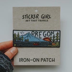 May include: Embroidered iron-on patch featuring an Oregon landscape with a mountain, trees, and a lake. The patch includes the word "OREGON" and the text "STICKER GIRL ART THAT TRAVELS". The patch is rectangular and attached to a cardboard backing.