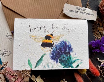 以下が含まれることがあります： 蜂と青い花が描かれた白いカード。カードには「happy bee-day!」と書かれており、裏には「植えてください、私は野花になります！」というメッセージが書かれています。