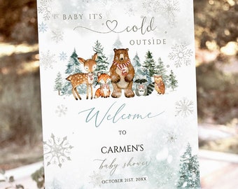 May include: A welcome sign for a baby shower, featuring a winter forest scene with woodland animals and snowflakes. The sign reads "Baby It's Cold Outside" and welcomes guests to Carmen's baby shower on October 21st.