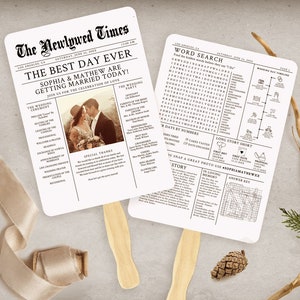 May include: Two white wedding fans with wooden handles, designed to look like newspapers. The headline reads "The Newlywed Times" and "The Best Day Ever." One fan features a photo of a couple, the other a word search. A beige ribbon, pinecone, and greenery are also present.