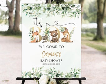 May include: A welcome sign for a baby shower with a woodland theme. The sign is white with green leaves and gold lettering. It says "It's a Boy" with a heart and a fox, raccoon, bear, and deer. The sign also says "Welcome to Carmen's Baby Shower October 21st, 20XX".