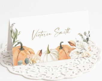 May include: A folded white place card with watercolor pumpkins and foliage. The card features the name "Victoria Smith" in gold script. The bottom right corner of the card has the text "BEINGHAPPYPRINTS".