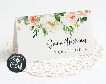 May include: White and pink floral wedding table card with the text "Sara Thomas" and "Table Three" in black script font.