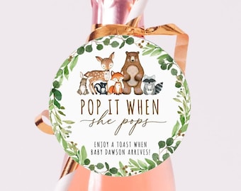 Puede incluir: Una pequeña botella de champán rosado con una pajita dorada y una pegatina redonda que dice "Pop it when she pops. Enjoy a toast when baby Dawson arrives!" La pegatina tiene una corona verde con animales del bosque, incluyendo un oso, un zorro, un ciervo y un mapache.