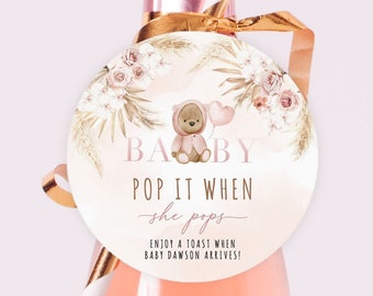 Puede incluir: Una etiqueta de favor de baby shower rosa y dorada con una ilustración de un osito de peluche y el texto "BABY POP IT WHEN she pops ENJOY A TOAST WHEN BABY DAWSON ARRIVES!"