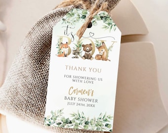 Op de afbeelding: Een wit label met groene bladeren en bosdieren die "het is een jongen" spellen in gouden letters. Het label is bevestigd aan een jute cadeauzak. De tekst op het label luidt "Bedankt voor het vieren van onze baby shower Carmen's Baby Shower July 24th, 20XX BeingHappyPrints".