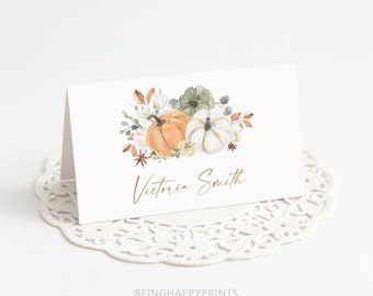 May include: A folded white card with a watercolor design of pumpkins and foliage in shades of orange, green, and white. The name "Victoria Smith" is written in gold script. The card is on a white lace doily.