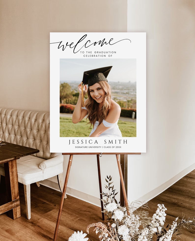 May include: White welcome sign with black calligraphy text that reads "Welcome to the Graduation Celebration of Jessica Smith Signature University Class of 20XX". A photo of a woman wearing a graduation cap and gown is in the center of the sign.