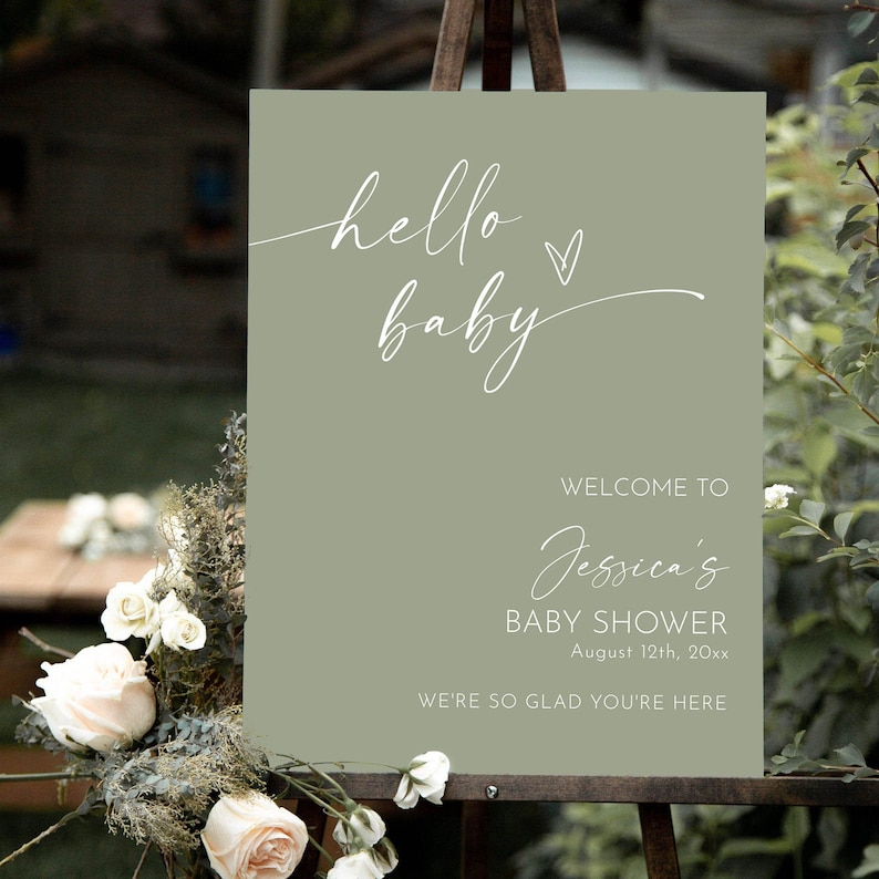 May include: A sage green welcome sign with white calligraphy that reads "hello baby" with a small heart. The sign also says "Welcome to Jessica's Baby Shower August 12th, 20xx We're so glad you're here."
