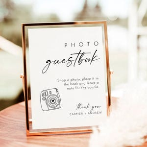 Pode incluir: Uma placa branca com moldura dourada, com o texto "PHOTO guestbook" em escrita cursiva preta. A placa instrui os convidados a "tirar uma foto, coloc&aacute;-la no livro e deixar uma nota para o casal". Abaixo do texto h&aacute; uma ilustra&ccedil;&atilde;o em preto e branco de uma c&acirc;mera instant&acirc;nea. A placa tamb&eacute;m inclui o texto "thank you CARMEN ANDREW".