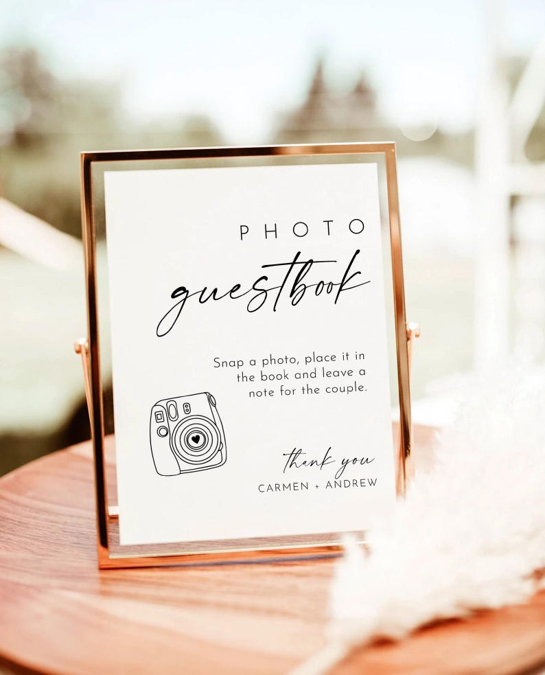 Pode incluir: Uma placa branca com moldura dourada, com o texto "PHOTO guestbook" em escrita cursiva preta. A placa instrui os convidados a "tirar uma foto, coloc&aacute;-la no livro e deixar uma nota para o casal". Abaixo do texto h&aacute; uma ilustra&ccedil;&atilde;o em preto e branco de uma c&acirc;mera instant&acirc;nea. A placa tamb&eacute;m inclui o texto "thank you CARMEN ANDREW".
