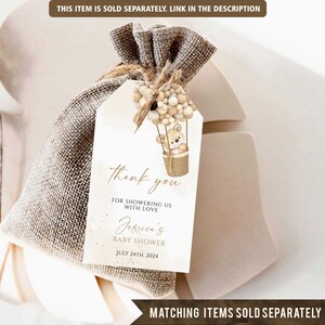 May include: A white thank you tag with a brown teddy bear in a hot air balloon with beige and brown balloons. The tag says "Thank you for showering us with love Jessica's Baby Shower July 24th, 2024".