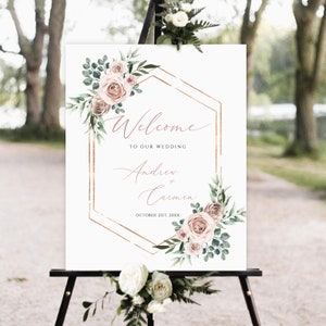 May include: A white wedding welcome sign with a gold geometric frame and pink floral accents. The text reads "Welcome to our wedding Andrew + Carmen October 21st, 20XX".