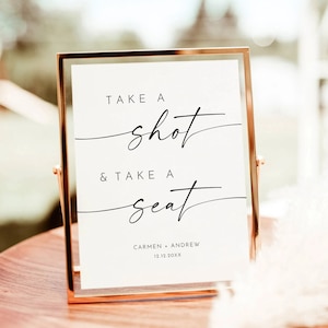 Puede incluir: Un cartel blanco en un marco dorado con el texto "Take a shot & take a seat" y los nombres Carmen y Andrew con la fecha 12.12.20XX.