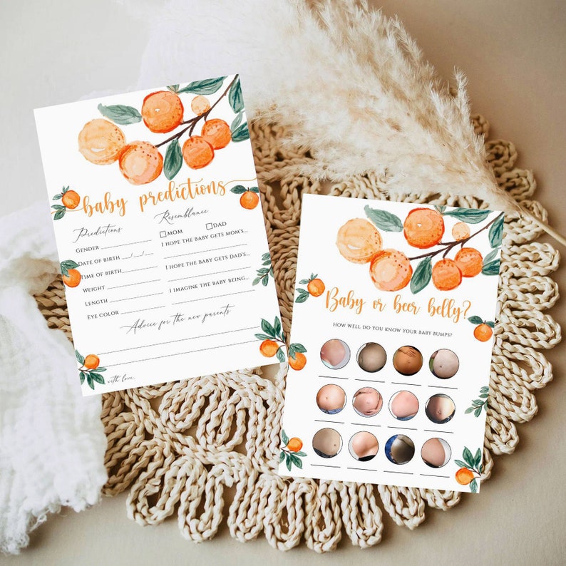 May include: Two baby shower game cards with watercolor orange and green leaf illustrations. One card reads "baby predictions" and the other says "Baby or beer belly?" with images of baby bumps. The cards are on a woven surface.