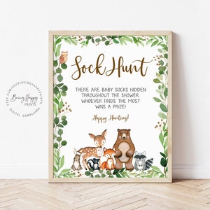 May include: A white sign with a brown border and green foliage around the edges. The sign says "Sock Hunt" in brown text. The text below says "There are baby socks hidden throughout the shower. Whoever finds the most wins a prize! Happy Hunting!" There are cartoon animals at the bottom of the sign, including a deer, a bear, a fox, a rabbit, a raccoon, and a skunk.