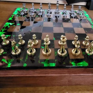 Puede incluir: Un juego de ajedrez con base de madera y borde de resina verde y negra. El tablero presenta piezas de ajedrez doradas y plateadas. Las piezas de ajedrez están dispuestas en el tablero a cuadros, listas para jugar.