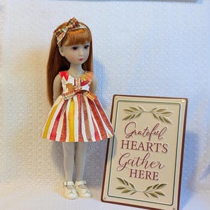 May include: A doll with red hair wearing a colorful dress with vertical stripes of red, yellow, and white. The doll has a matching headband and white shoes. A sign next to the doll reads "Grateful Hearts Gather HERE."