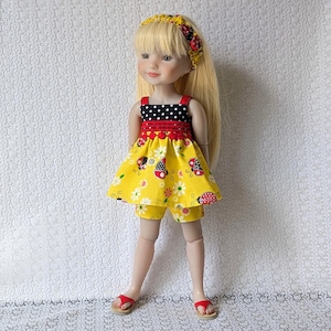 May include: A doll wearing a yellow ladybug-themed outfit, including a dress and shorts. The dress has a black and white polka dot top with red trim and a yellow skirt with ladybug and flower patterns. The doll also wears a matching headband and red sandals.