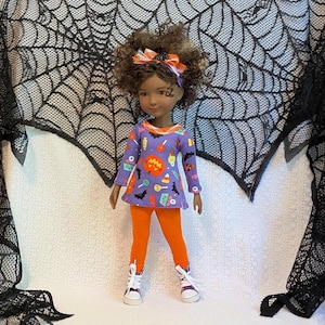 Könnte beinhalten: Eine Puppe in einer lila Halloween-Tunika, orangefarbenen Leggings und weißen Turnschuhen. Die Tunika zeigt einen Kürbis, Fledermäuse und andere Halloween-Symbole. Die Puppe hat lockiges braunes Haar und eine orange-weißes Schleife.