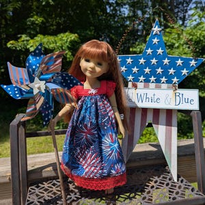 May include: A doll with long auburn hair wearing a navy blue dress with a red, white, and blue firework pattern and red lace trim. The doll holds a patriotic pinwheel with stars and stripes. Behind the doll is a metal star sign with the text Red, White & Blue.