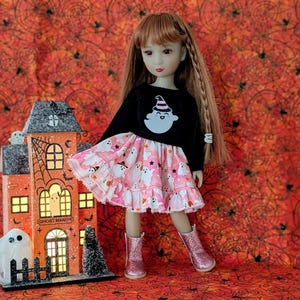 May include: A doll with long brown hair wearing a black long-sleeve shirt with a white ghost graphic and a pink skirt featuring a ghost pattern. The doll wears glittery pink boots and stands next to a light-up orange house labeled Ghost Manor.