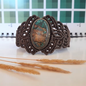 May include: A brown macrame bracelet with a large oval-shaped turquoise gemstone set in a brass bezel. The bracelet is on a white surface with dried grass.