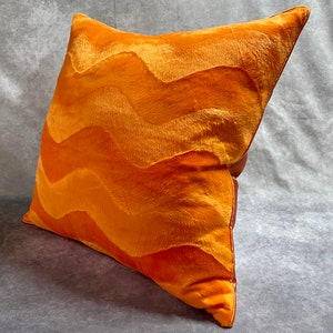 K&ouml;nnte beinhalten: Orangefarbenes Samtkissen mit einem wellenf&ouml;rmigen Muster. Das Kissen ist aus einem weichen, pl&uuml;schigen Stoff gefertigt und hat einen dezenten Glanz.