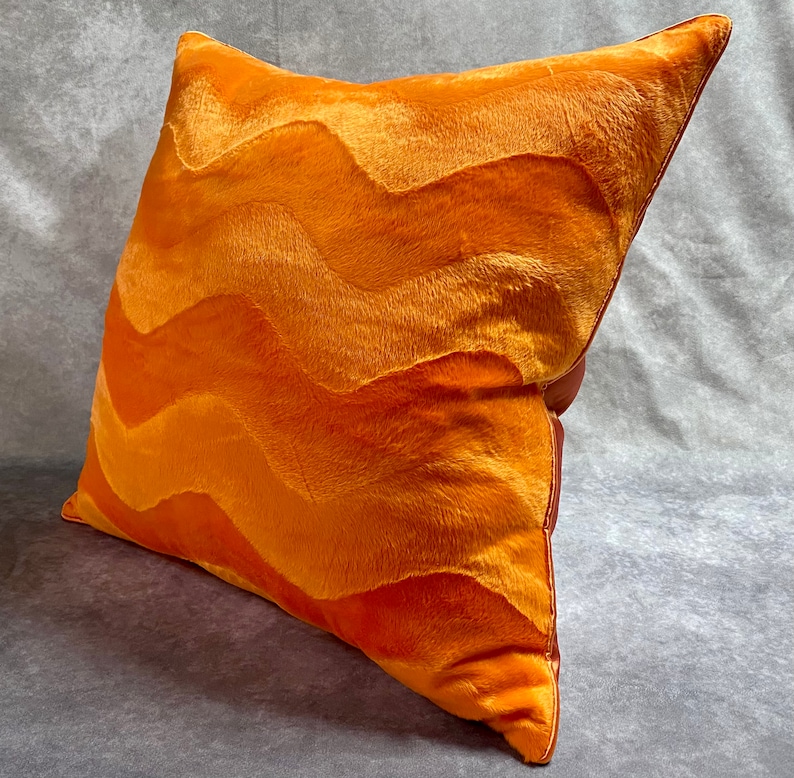 K&ouml;nnte beinhalten: Orangefarbenes Samtkissen mit einem wellenf&ouml;rmigen Muster. Das Kissen ist aus einem weichen, pl&uuml;schigen Stoff gefertigt und hat einen dezenten Glanz.