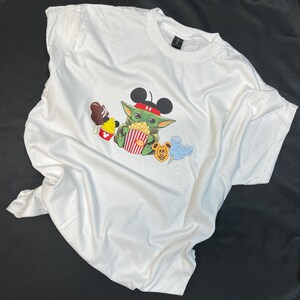 May include: White t-shirt with a green character wearing Mickey Mouse ears and holding a bucket of popcorn. The character is surrounded by other Disney characters, including Mickey Mouse, a blue ice cream cone, and a brown ice cream bar.