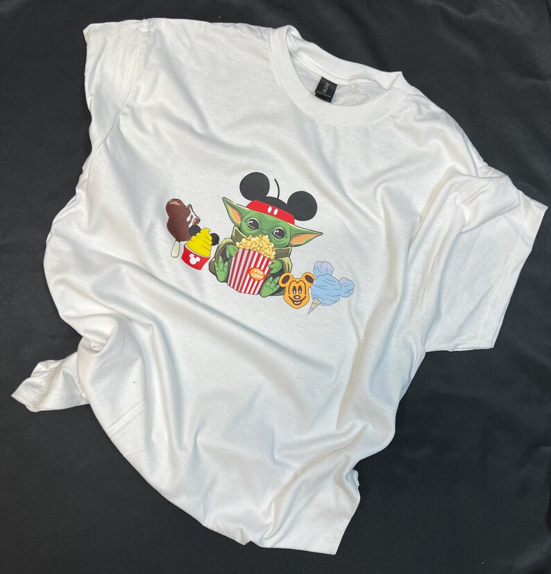 May include: White t-shirt with a green character wearing Mickey Mouse ears and holding a bucket of popcorn. The character is surrounded by other Disney characters, including Mickey Mouse, a blue ice cream cone, and a brown ice cream bar.