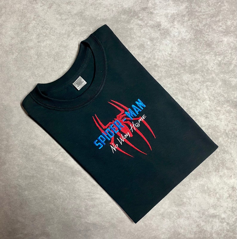 以下が含まれることがあります： 赤い糸と青い糸で刺繍されたスパイダーマンのロゴと「ノー・ウェイ・ホーム」の文字が書かれた黒いTシャツ。