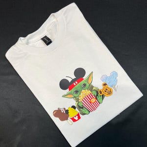 May include: White t-shirt with a green character wearing Mickey Mouse ears and holding a red and white striped popcorn bucket. The character is surrounded by other Disney-themed snacks, including a Mickey Mouse ice cream cone, a chocolate-covered Mickey Mouse ear, and a blue cotton candy.