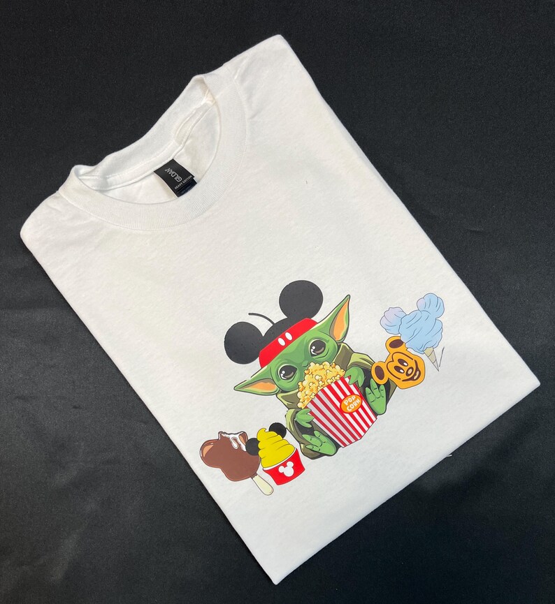May include: White t-shirt with a green character wearing Mickey Mouse ears and holding a red and white striped popcorn bucket. The character is surrounded by other Disney-themed snacks, including a Mickey Mouse ice cream cone, a chocolate-covered Mickey Mouse ear, and a blue cotton candy.