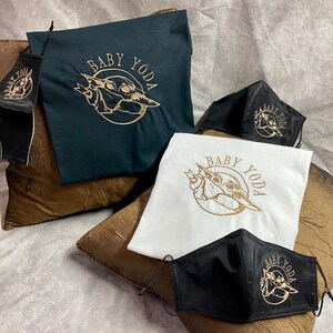 May include: A set of three black face masks and two t-shirts featuring a gold embroidered image of Baby Yoda. The t-shirts are one black and one white. The masks and t-shirts all have the text "Baby Yoda" embroidered on them.