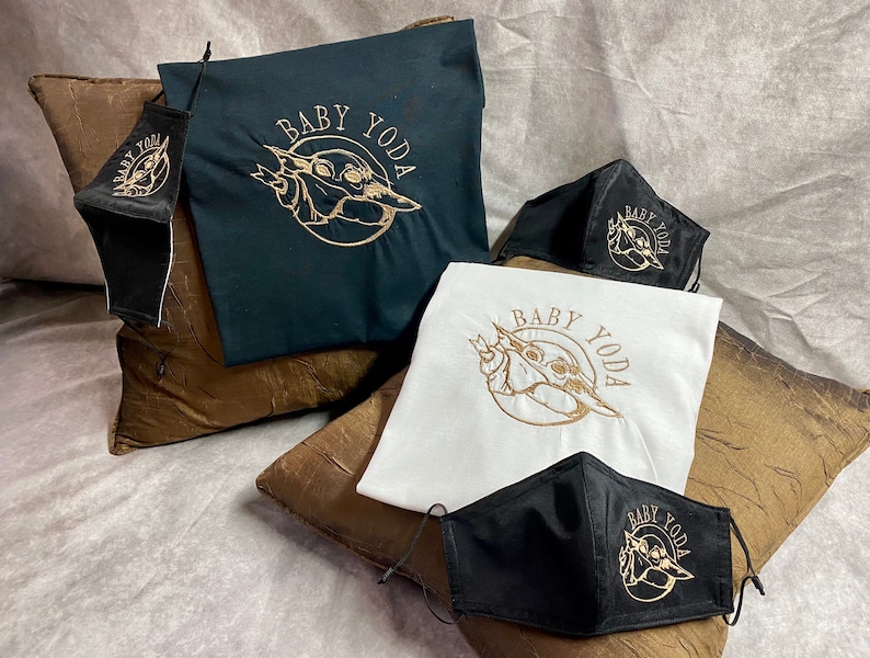 May include: A set of three black face masks and two t-shirts featuring a gold embroidered image of Baby Yoda. The t-shirts are one black and one white. The masks and t-shirts all have the text "Baby Yoda" embroidered on them.