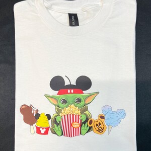 May include: White t-shirt with a green character wearing Mickey Mouse ears and a red headband, holding a red and white striped popcorn bucket. The character is surrounded by Mickey Mouse-themed snacks, including a Mickey Mouse ice cream cone, a Mickey Mouse ice cream sandwich, and a Mickey Mouse cotton candy.
