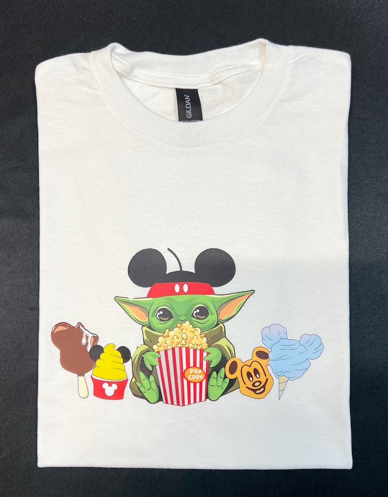 May include: White t-shirt with a green character wearing Mickey Mouse ears and a red headband, holding a red and white striped popcorn bucket. The character is surrounded by Mickey Mouse-themed snacks, including a Mickey Mouse ice cream cone, a Mickey Mouse ice cream sandwich, and a Mickey Mouse cotton candy.