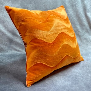 K&ouml;nnte beinhalten: Ein pl&uuml;schiges orangefarbenes Kissen mit einem wellenf&ouml;rmigen Muster. Das Kissen ist aus einem weichen, flauschigen Stoff gefertigt und hat einen dezenten Glanz.