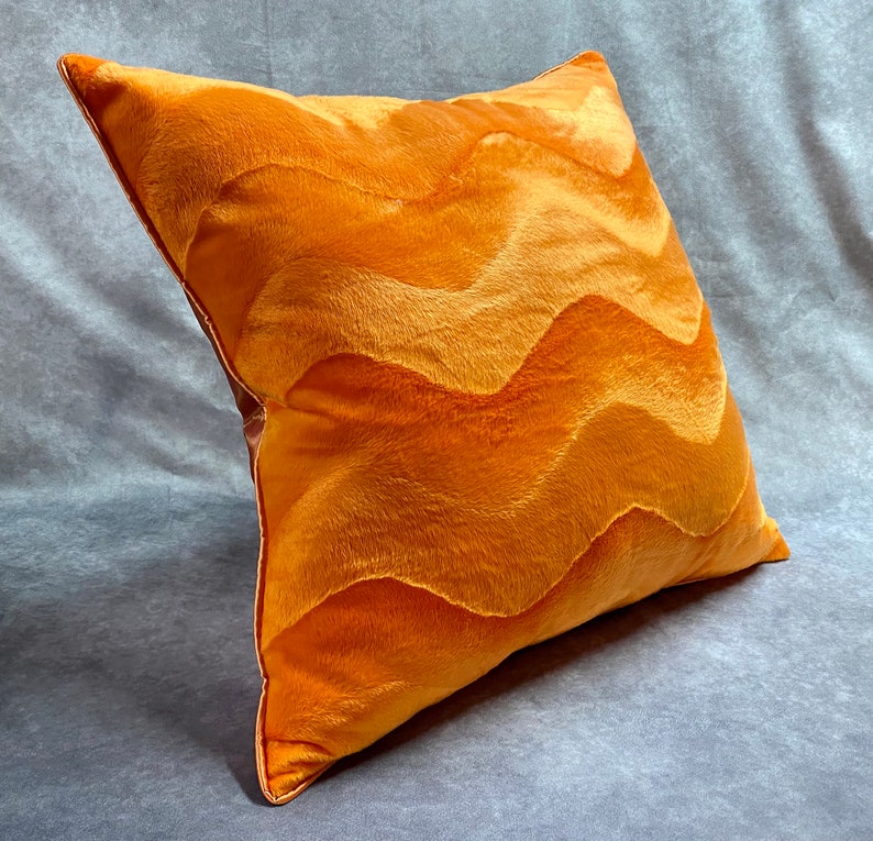 K&ouml;nnte beinhalten: Ein pl&uuml;schiges orangefarbenes Kissen mit einem wellenf&ouml;rmigen Muster. Das Kissen ist aus einem weichen, flauschigen Stoff gefertigt und hat einen dezenten Glanz.