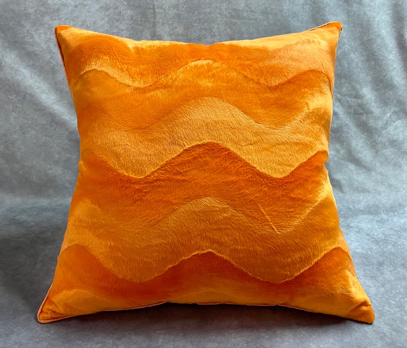 K&ouml;nnte beinhalten: Ein pl&uuml;schiges orangefarbenes Kissen mit einem wellenf&ouml;rmigen Muster. Das Kissen ist aus einem weichen, flauschigen Material gefertigt und hat einen dezenten Glanz.