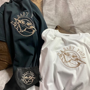 May include: Two t-shirts with a gold embroidered image of Baby Yoda. The text "Baby Yoda" is embroidered in gold on a white t-shirt. The text "Baby Y" is embroidered in gold on a black t-shirt.