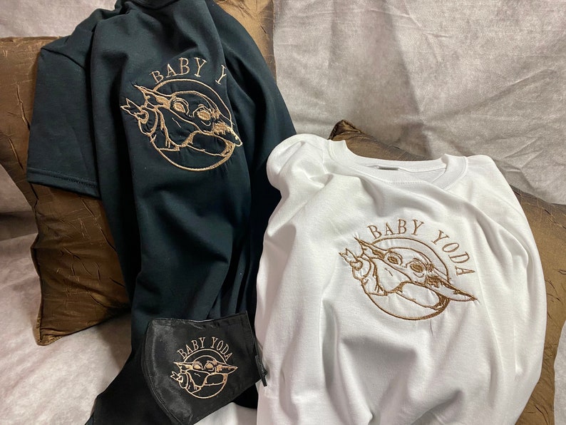 May include: Two t-shirts with a gold embroidered image of Baby Yoda. The text "Baby Yoda" is embroidered in gold on a white t-shirt. The text "Baby Y" is embroidered in gold on a black t-shirt.