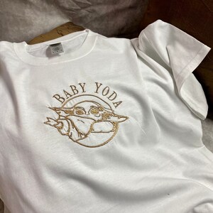 May include: White t-shirt with a gold embroidered design of Baby Yoda. The design includes the text "Baby Yoda" in a circle around the character.