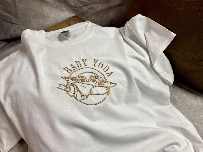 May include: White t-shirt with a gold embroidered design of Baby Yoda. The design includes the text "Baby Yoda" in a circle around the character.