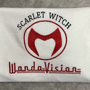 May include: White t-shirt with red embroidered text "Scarlet Witch" and a red "M" symbol in a circle. Below the symbol is the text "WandaVision" in red.
