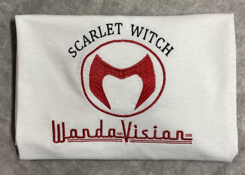 May include: White t-shirt with red embroidered text "Scarlet Witch" and a red "M" symbol in a circle. Below the symbol is the text "WandaVision" in red.