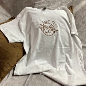 May include: White t-shirt with a brown embroidered image of Baby Yoda and the text "Baby Yoda".
